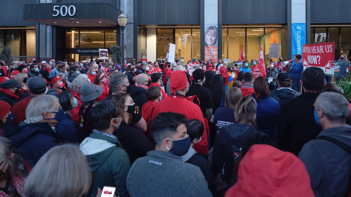 A dense crowd of healthcare workers, predominantly wearing red jackets and face masks, congregates outside the Kaiser Permanente Tower at 500 Northeast Multnomah Street in Portland's Lloyd District. The sea of protesters fills the frame from foreground to the building's glass facade, creating a powerful visual of collective action. Protest signs punctuate the crowd, including one reading 'CAN YOU HEAR US NOW?' while the building's modernist architecture looms behind them, its illuminated interior contrasting with the gathering dusk outside.