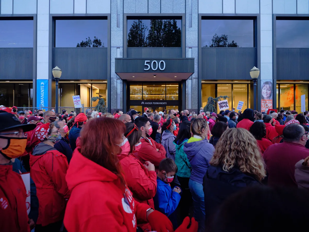 Hundreds of nurses in vibrant red union jackets and winter coats converge in the twilight hours outside the Kaiser Permanente Tower on Northeast Multnomah Street in Portland's Lloyd District. The crowd creates a sea of crimson against the modern glass facade of the 500 building, their collective presence illuminated by the warm glow spilling from the medical facility's entrance. The atmospheric blue hour lighting captures the gravity and determination of healthcare workers preparing for labor action, with protest signs visible among the densely packed crowd.