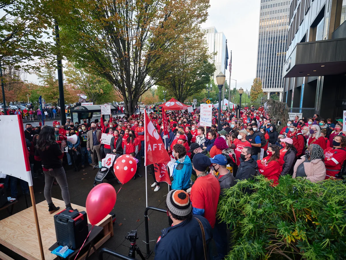 Hundreds of nurses and healthcare workers in vibrant red union attire congregate outside Kaiser Permanente's towering glass headquarters in Portland's Lloyd District, their determined faces reflecting the gravity of impending labor action. The autumn foliage of street trees creates a golden canopy above the crowd, while red AFT banners and protest signs punctuate the sea of solidarity. A makeshift speaking platform commands attention from the diverse assembly, capturing the pivotal moment before a significant healthcare strike unfolds against the urban backdrop of downtown Portland.