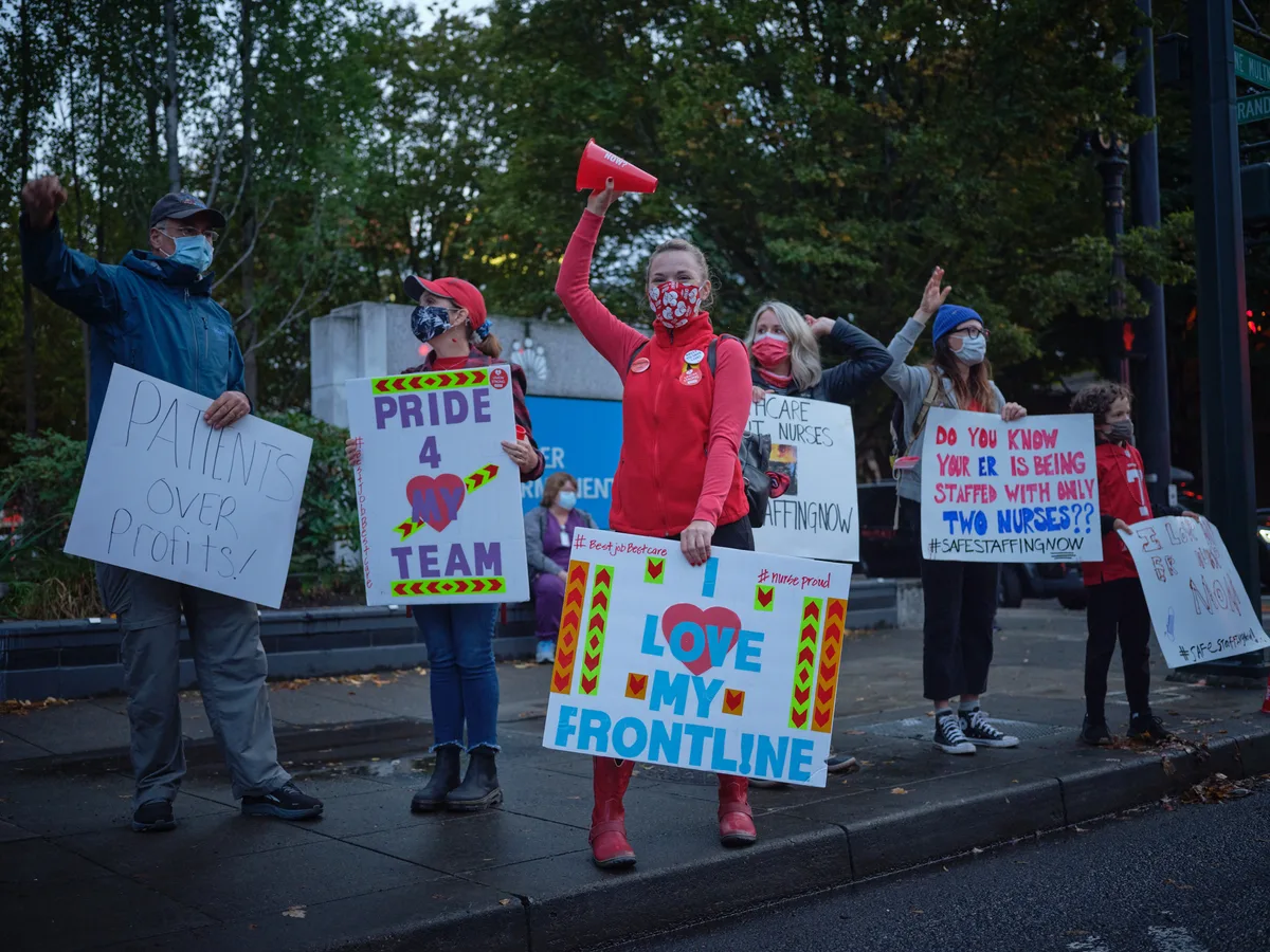 Healthcare workers gather on the sidewalk outside Kaiser Permanente Tower in Portland's Lloyd District, their voices unified in protest against inadequate staffing levels. Masked demonstrators hold colorful handmade signs declaring 'Love My Frontline' and demanding safe patient-to-nurse ratios, their red scrubs and bright placards creating vivid punctuation marks against the muted autumn evening. The golden hour light filters through towering street trees, casting long shadows across the concrete as nurses raise their fists and megaphones in solidarity. The scene captures the determined spirit of frontline workers advocating for both patient safety and workplace dignity during a pivotal labor action.