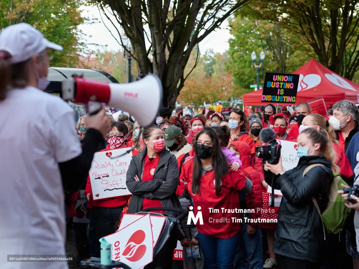 A sea of red-clad Kaiser Permanente nurses and healthcare workers mass beneath autumn trees in Portland, their unified presence creating a powerful visual statement of solidarity. A strike organizer commands attention through a megaphone in the foreground, her white baseball cap and focused stance contrasting against the dense crowd of masked protesters behind her. Handmade signs demanding "Quality Care" and union representation punctuate the gathering, while the warm fall light filters through changing leaves, casting an almost pastoral backdrop to this moment of labor activism.