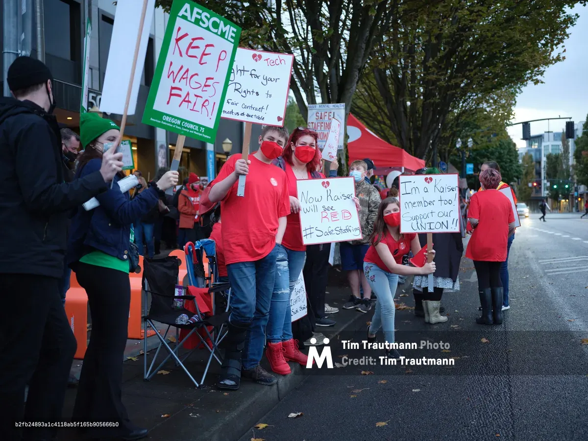 Healthcare workers in crimson masks and shirts gather on Northeast Multnomah Street outside Kaiser Permanente Tower, their handmade signs demanding fair wages and union recognition piercing the overcast Portland evening. The AFSCME banners and personal placards create a sea of red against the urban backdrop, while folding chairs and determined faces signal a prolonged demonstration. Autumn leaves scatter across wet pavement as protesters maintain their vigil beneath the towering medical complex, their collective voice rising in the Lloyd District's fading light.