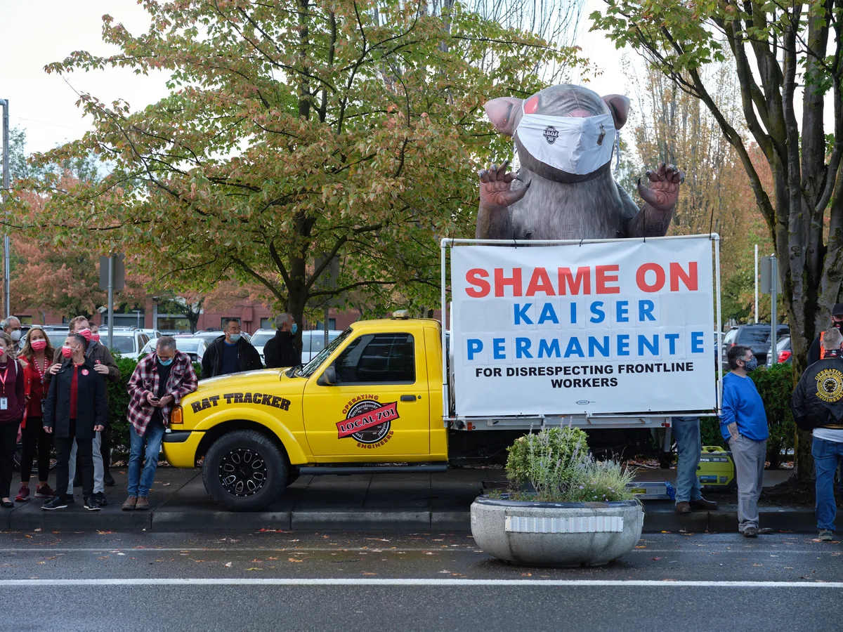 A towering inflatable rat wearing a surgical mask looms over a labor demonstration in Portland's Lloyd District, its claws raised menacingly beside a bright yellow truck bearing the message 'Shame on Kaiser Permanente.' Masked healthcare workers and union supporters cluster around the symbolic creature beneath autumn trees, their presence creating a powerful tableau of organized dissent. The overcast Oregon sky provides a muted backdrop to the bold red and blue protest signage, while fallen leaves scattered across the pavement hint at the season of change both literally and figuratively.