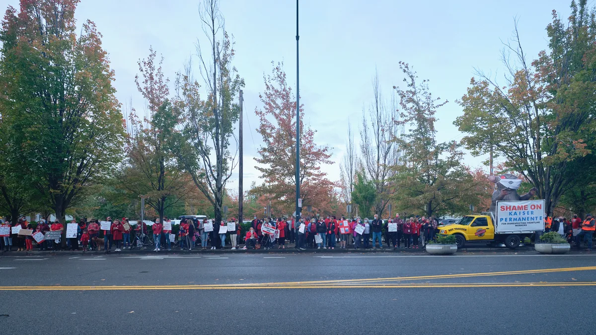 A sea of red-clad nurses and supporters mass along Northeast Multnomah Street outside Kaiser Permanente Tower in Portland's Lloyd District, their protest signs creating a rhythmic pattern against autumn's burnished foliage. The overcast Pacific Northwest sky casts diffused light across the demonstration, while a yellow union truck bearing "SHAME ON KAISER PERMANENTE" anchors the right side of the frame. The striking healthcare workers stretch in an unbroken line along the sidewalk, their collective presence transforming the corporate streetscape into a theater of labor solidarity.