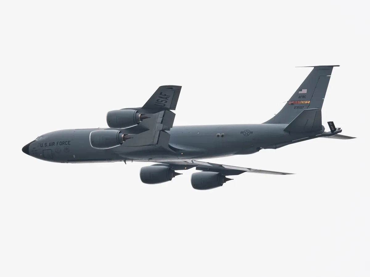 A U.S. Air Force Boeing KC-135 Stratotanker aerial refueling aircraft photographed in flight from below against a flat, overcast white sky. The aircraft is painted in overall grey and bears "U.S. AIR FORCE" lettering on the fuselage. "USAF" is stenciled on the vertical stabilizer, with "AFRC" and "MARCH" unit markings and the tail number 23507 visible near the tail. Four podded jet engines are mounted under the swept wings. The aircraft's boom refueling equipment is visible extending from the rear of the fuselage. A U.S. national insignia roundel appears on the fuselage. The tight telephoto framing isolates the aircraft cleanly against the grey-white sky.