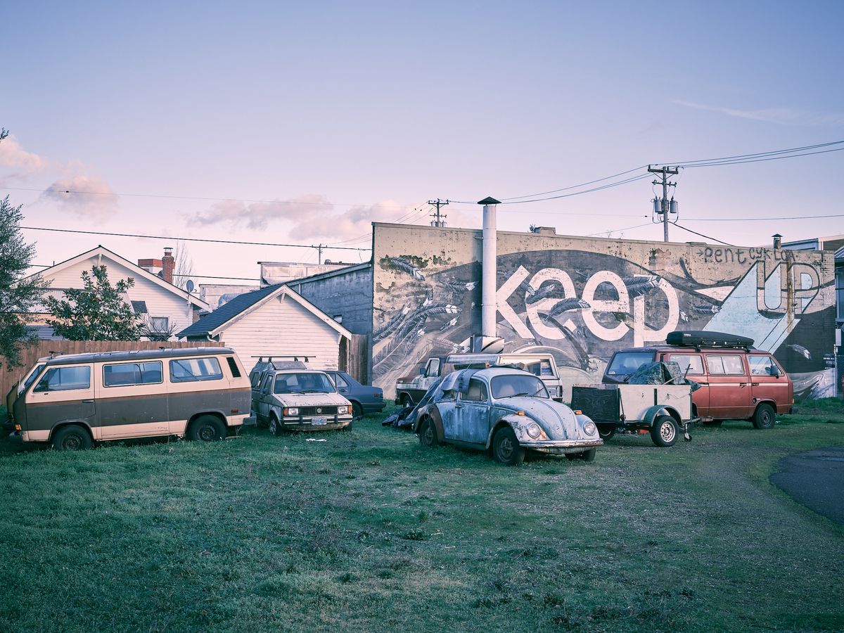 A collection of aging and worn vehicles, including two Volkswagen Vanagons, a rusted VW Beetle, and a red van with a roof rack, are parked on a grassy lot in Portland's Alberta Arts District. Behind them, a large weathered building displays a partial mural reading "Keep...Up," part of the "Keep Your Chin Up" street art piece, featuring painted feathers and organic forms in black, white, and blue. Residential homes, utility poles, and a soft purple-hued evening sky are visible in the background.