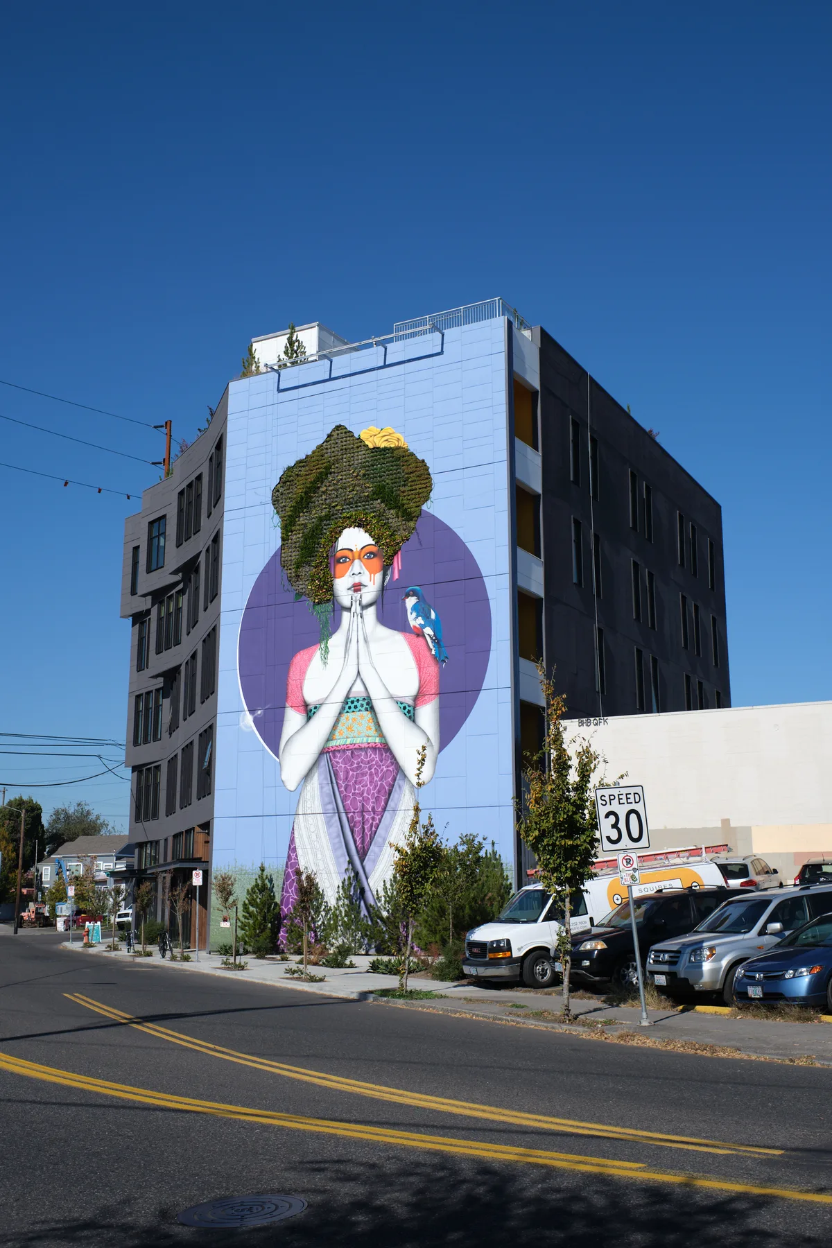 Against Portland's crystalline azure sky, Fin DAC's monumental five-story mural transforms a contemporary apartment building into a beacon of contemplative art. The ethereal figure, crowned with cascading vegetation that breathes life into street art, presses palms together in prayer while a vibrant parrot perches nearby, creating an oasis of serenity amid the urban pulse of Southeast Division Street. Yellow lane markers slice through the foreground, leading the eye toward this towering testament to gratitude that seamlessly merges nature with human expression.