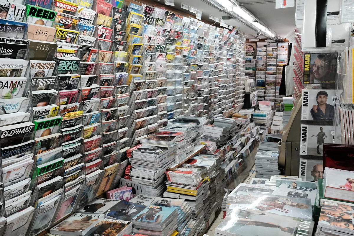 The cramped interior of a traditional newsstand on Prince Street in SoHo reveals an overwhelming kaleidoscope of publications. Floor-to-ceiling magazine racks create narrow corridors lined with hundreds of titles, from fashion glossies to niche periodicals, while harsh fluorescent lighting illuminates the densely packed space. Stacks of fresh magazines and newspapers sprawl across every available surface, creating a labyrinthine environment that speaks to the enduring appetite for print media in Manhattan's cultural heart.