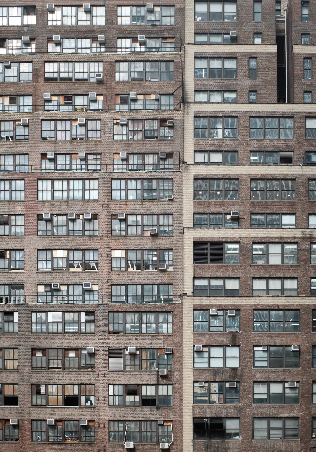 A dense pattern of weathered brick apartment building facades creates an urban tapestry in Midtown Manhattan. The repetitive grid of windows, punctuated by air conditioning units and varying states of wear, reveals the intimate details of city living through glimpses of curtains, blinds, and personal belongings. The warm brown and red brick tones contrast with the cool reflective glass, while the geometric composition emphasizes the vertical density of New York residential architecture.
