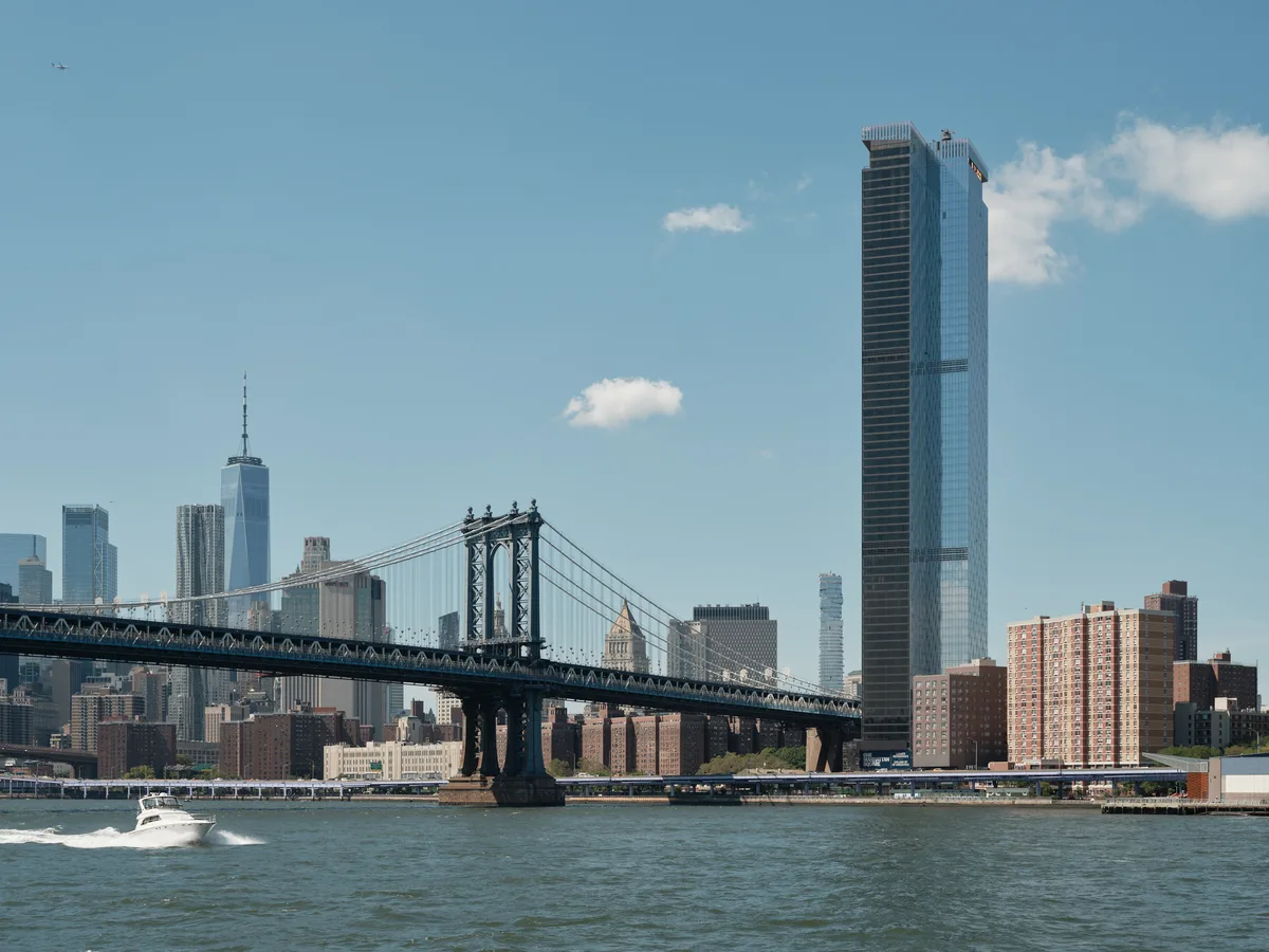 The iconic Manhattan Bridge stretches across the East River in elegant suspension, its steel towers and cables creating geometric lines against a pale blue afternoon sky dotted with cotton-white clouds. One World Trade Center's gleaming spire punctuates the left side of Manhattan's financial district, while a striking dark glass tower dominates the right portion of the frame. A white pleasure boat cuts through the choppy river waters in the foreground, leaving a gentle wake as it passes beneath this architectural symphony of old and new New York.