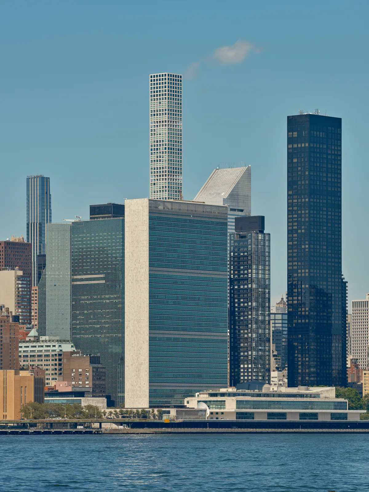 The iconic Manhattan skyline rises majestically from the East River, captured from Brooklyn's Greenpoint neighborhood on a crystalline afternoon. The United Nations Headquarters' distinctive rectangular form anchors the composition alongside gleaming residential towers, their glass facades catching the clear light against an azure sky. Gentle ripples disturb the river's surface in the foreground, while wispy clouds drift across the expansive urban panorama. The interplay of modernist architecture and classical skyscraper forms creates a compelling study of New York's evolving vertical landscape.
