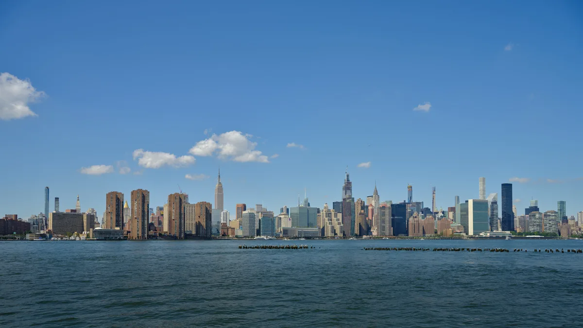 The iconic Manhattan skyline rises majestically across the East River, viewed from the industrial waterfront of Greenpoint, Brooklyn. Wispy white clouds drift across an azure afternoon sky, creating a serene backdrop for the dense cluster of skyscrapers including the distinctive Empire State Building. The foreground features weathered wooden pier pilings emerging from the dark river waters, adding rustic texture to this classic New York vista. The composition captures the dramatic contrast between Brooklyn's quieter shores and Manhattan's towering urban density.