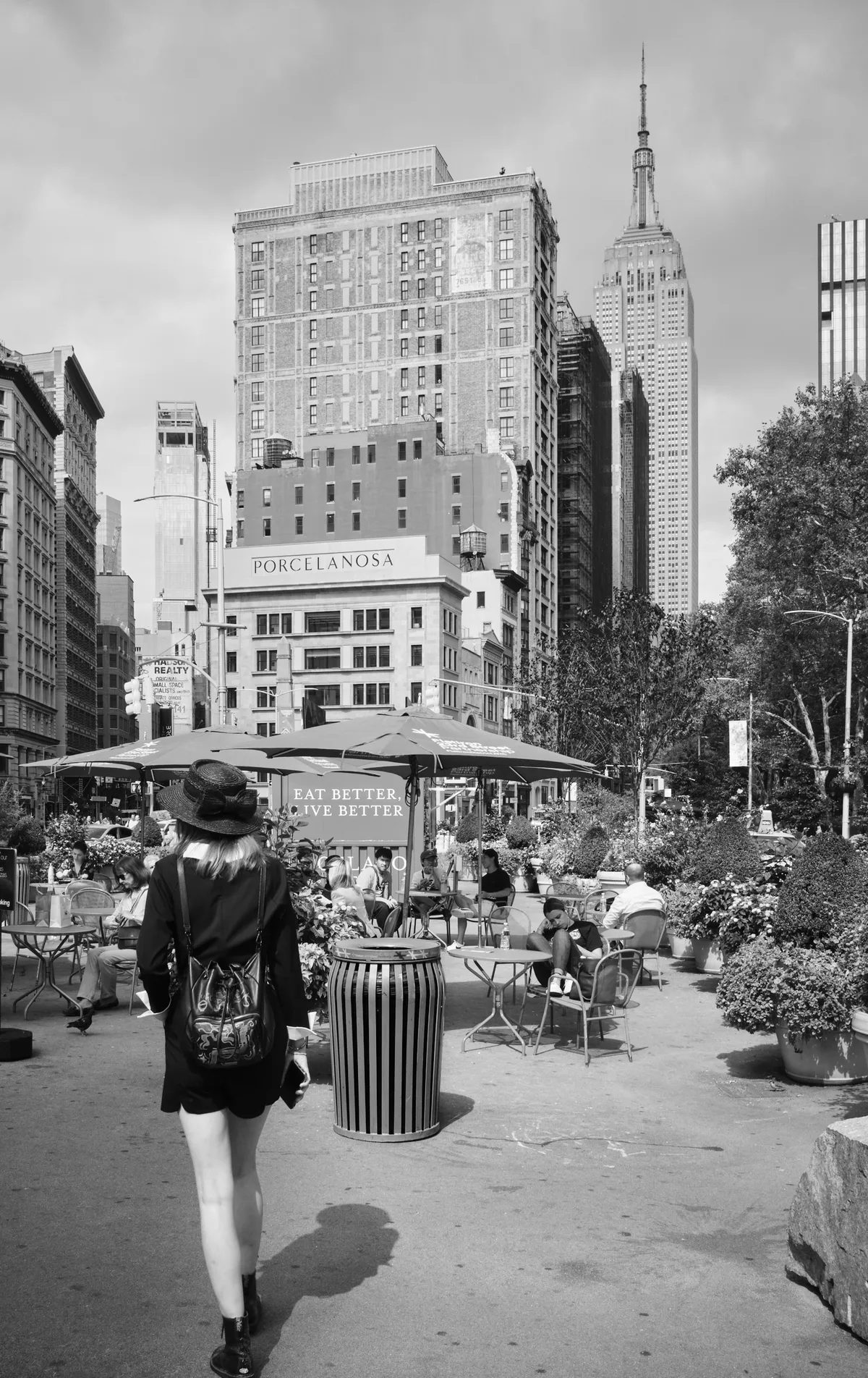A contemplative woman in a wide-brimmed hat pauses at the edge of Madison Square Park's bustling outdoor dining area, her shadow stretching across sun-warmed concrete. The Empire State Building rises majestically in the background, its Art Deco spire piercing overcast skies, while the historic Porcelanosa building and contemporary high-rises create Manhattan's signature architectural layering. The scene captures the gentle rhythm of urban life, where nature and commerce intersect in temporary harmony beneath market umbrellas and flowering planters.