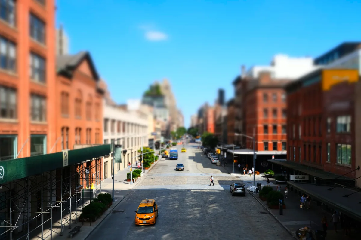 Shot from the elevated perspective of Manhattan's High Line park, this tilt-shift photograph transforms a bustling city street into a dreamlike miniature world. The shallow depth of field blurs the historic brick warehouses of the Meatpacking District into soft coral and burgundy tones, while a solitary yellow taxi cab sits in sharp focus on the sun-drenched asphalt below. The azure sky stretches endlessly above the urban canyon, creating a striking contrast with the warm industrial architecture that defines this transformed neighborhood.