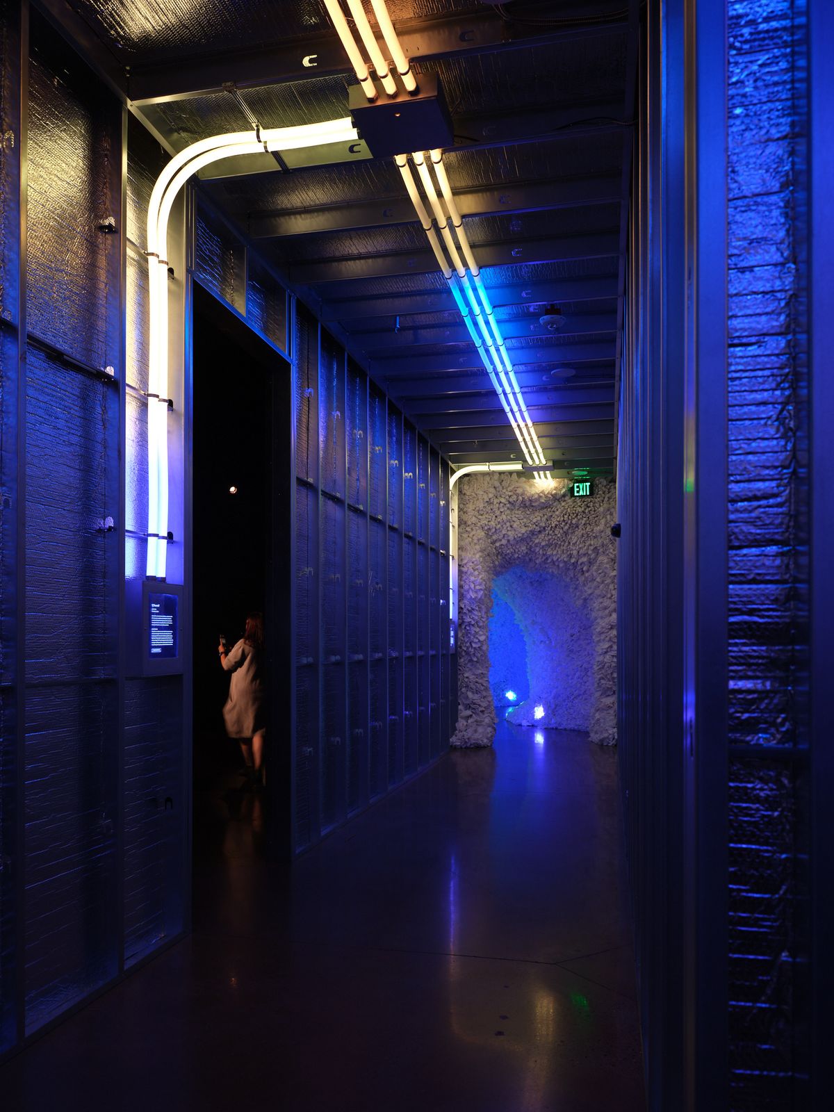 A dramatic interior hallway featuring blue LED lighting, exposed brick walls, and modern architectural elements including curved white light fixtures overhead. A person in light clothing stands in the corridor, emphasizing the scale of the space.