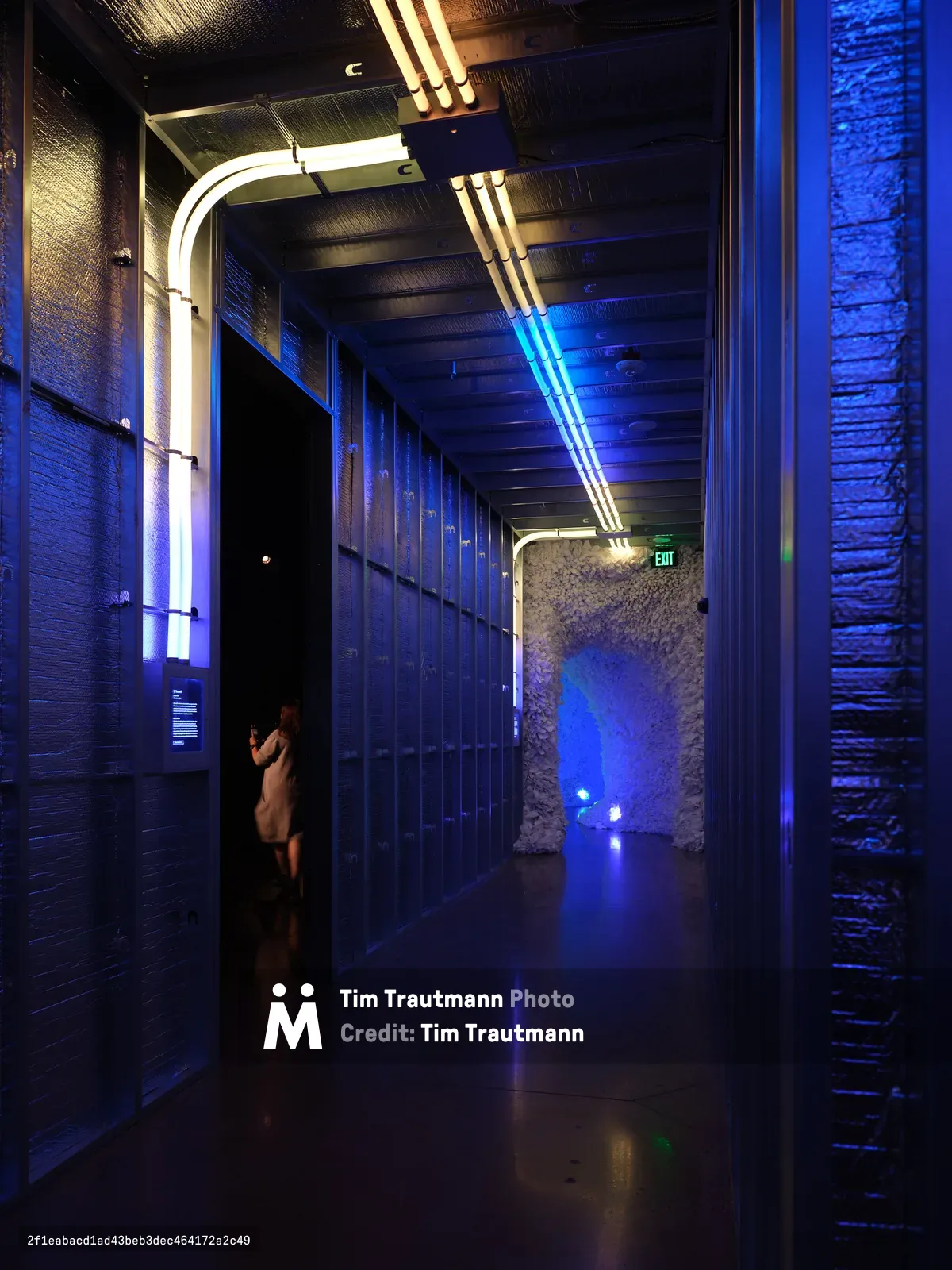 A dramatic interior hallway featuring blue LED lighting, exposed brick walls, and modern architectural elements including curved white light fixtures overhead. A person in light clothing stands in the corridor, emphasizing the scale of the space.