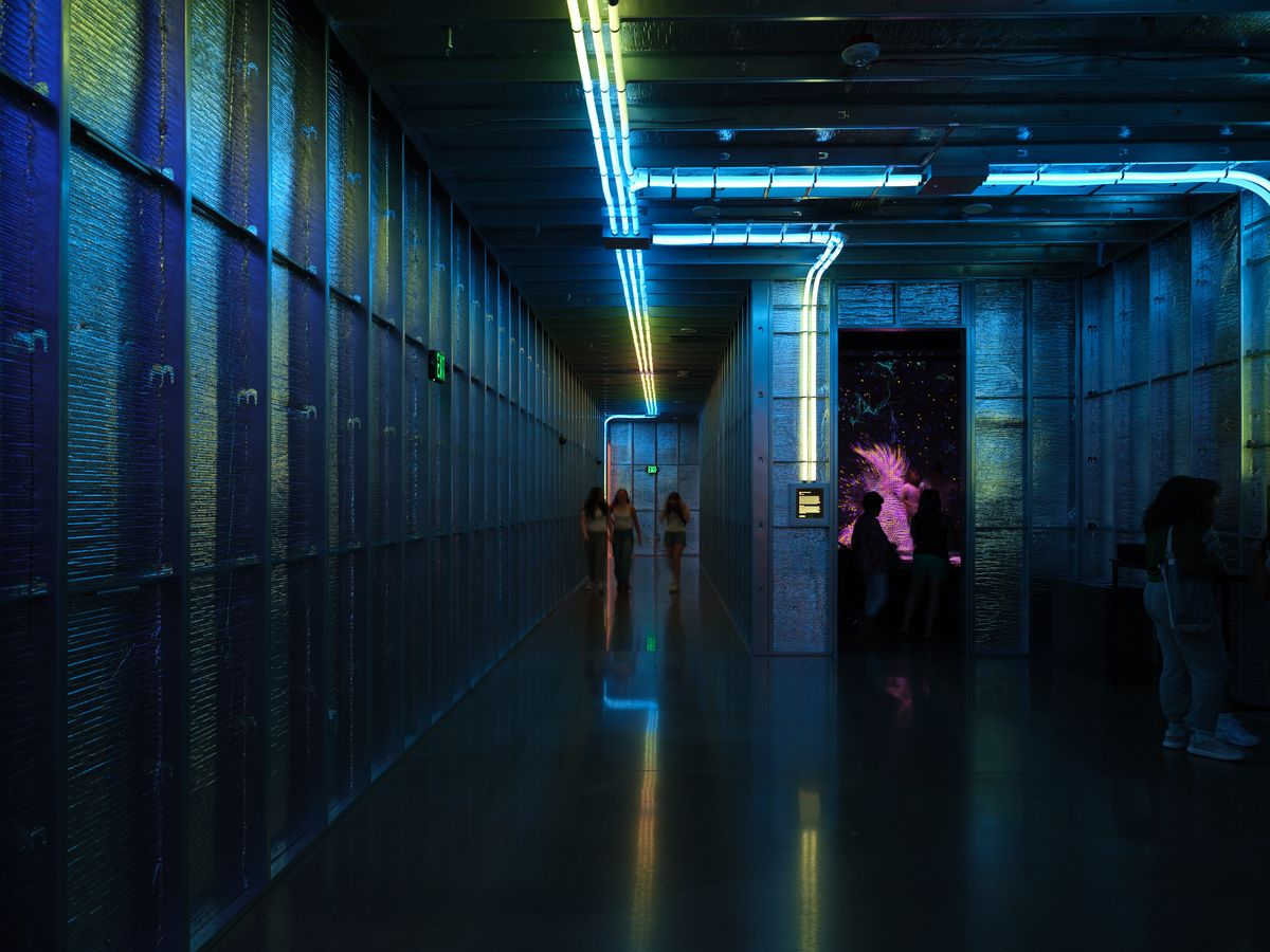 A futuristic museum hallway at Hopscotch in Portland, Oregon features dramatic blue neon lighting strips along the ceiling and walls, creating atmospheric illumination for visitors exploring the contemporary art space. The industrial architecture with exposed brick walls and polished floors reflects the vibrant lighting while silhouetted figures move through the corridor.