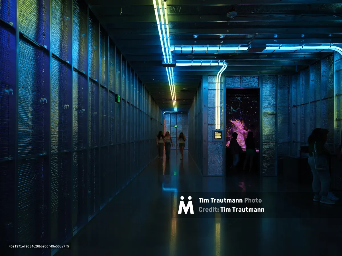 A futuristic museum hallway at Hopscotch in Portland, Oregon features dramatic blue neon lighting strips along the ceiling and walls, creating atmospheric illumination for visitors exploring the contemporary art space. The industrial architecture with exposed brick walls and polished floors reflects the vibrant lighting while silhouetted figures move through the corridor.