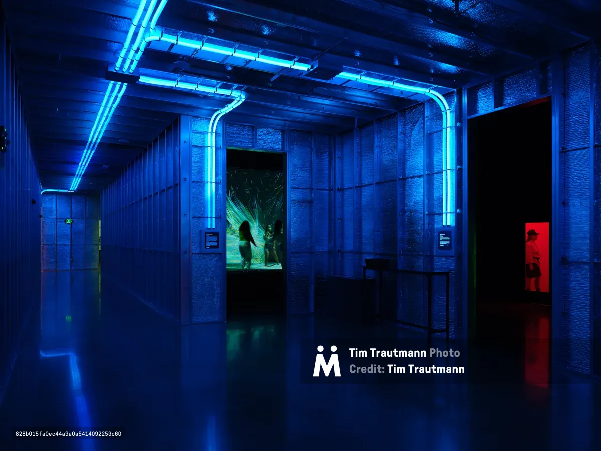 A contemporary museum gallery space in Portland featuring dramatic blue neon lighting strips along the ceiling and walls, with exhibition displays visible in the background and a striking red accent light on the right side. The polished concrete floor reflects the atmospheric lighting, creating a futuristic ambiance.