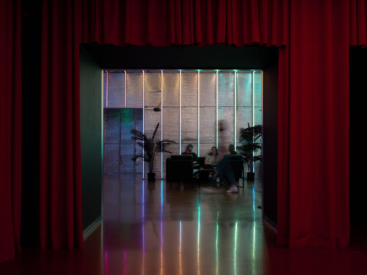 View through red curtains into a contemporary restaurant dining room featuring vertical neon light strips in rainbow colors along wood-paneled walls, with diners seated at tables and palm plants as decor. The polished floor reflects the vibrant lighting creating a modern, upscale atmosphere.