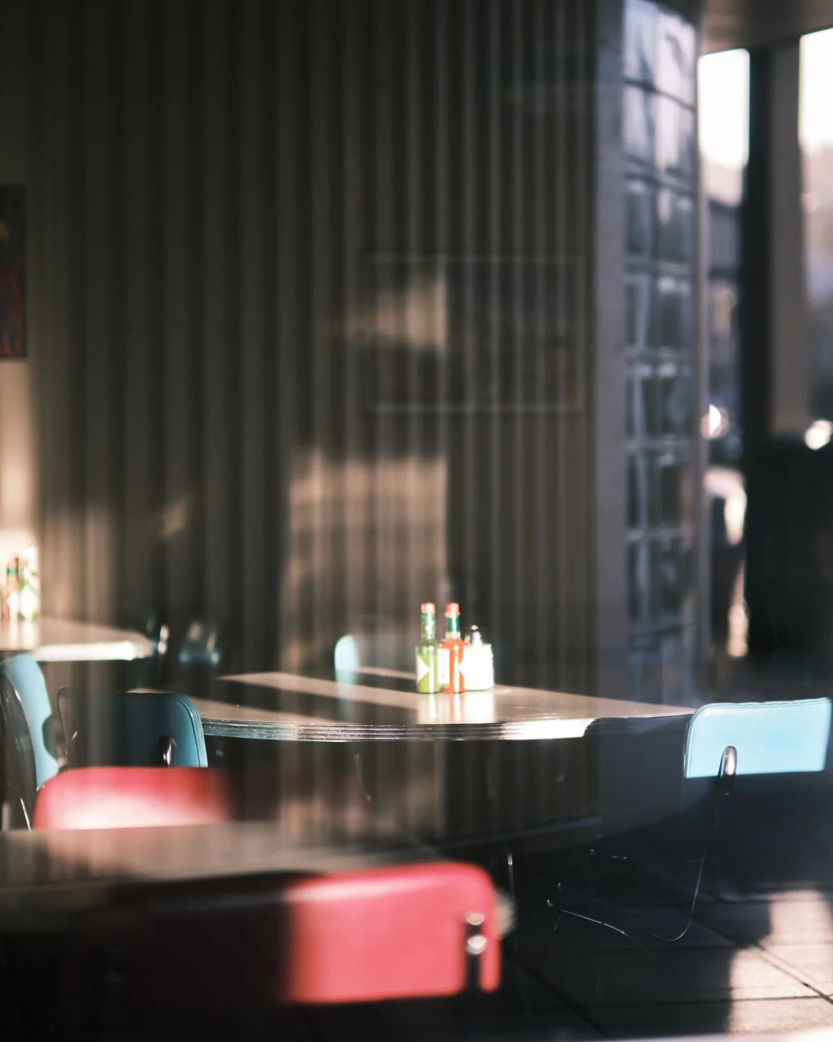 Soft morning light filters through tall windows, casting gentle shadows across a quiet corner table in a Seattle café. The scene captures the intimate atmosphere of urban breakfast culture, with condiment bottles centered on a round wooden table surrounded by red vinyl stools and light blue chairs. The shallow depth of field creates a dreamy quality, with the background dissolving into warm bokeh while the foreground maintains sharp detail on the simple table setting.