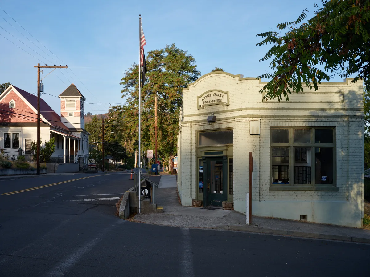 The historic Mosier Valley Post Office stands as a weathered sentinel along Oregon's quiet main thoroughfare, its cream-colored brick facade catching the warm embrace of late afternoon light. The Art Deco-influenced building, with its distinctive curved parapet and large storefront windows, anchors this Columbia River Gorge community while an American flag waves gently overhead. Residential homes with red roofs dot the tree-lined street beyond, creating an intimate portrait of small-town Pacific Northwest life where community institutions remain the heart of daily existence.
