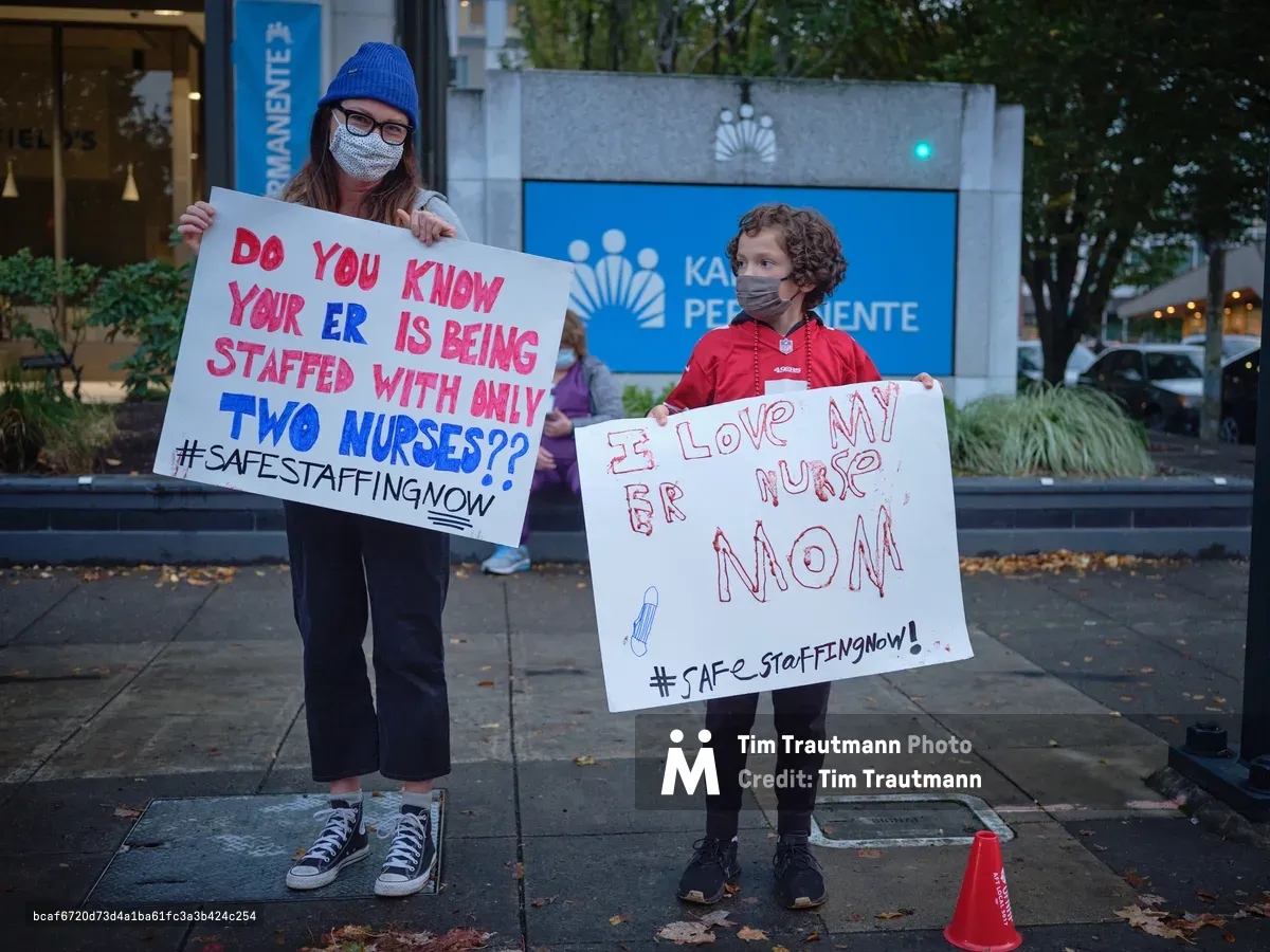 A woman in a cobalt blue knit beanie and face mask holds a handwritten protest sign questioning emergency room staffing levels, while a young child beside her displays a heartfelt message of support for their nurse mother. The pair stands on autumn-scattered sidewalks outside Kaiser Permanente's towering headquarters in Portland's Lloyd District, their white poster boards catching the soft urban light against the healthcare giant's signature blue corporate signage. The intimate scale of their grassroots demonstration contrasts powerfully with the institutional architecture looming behind them.