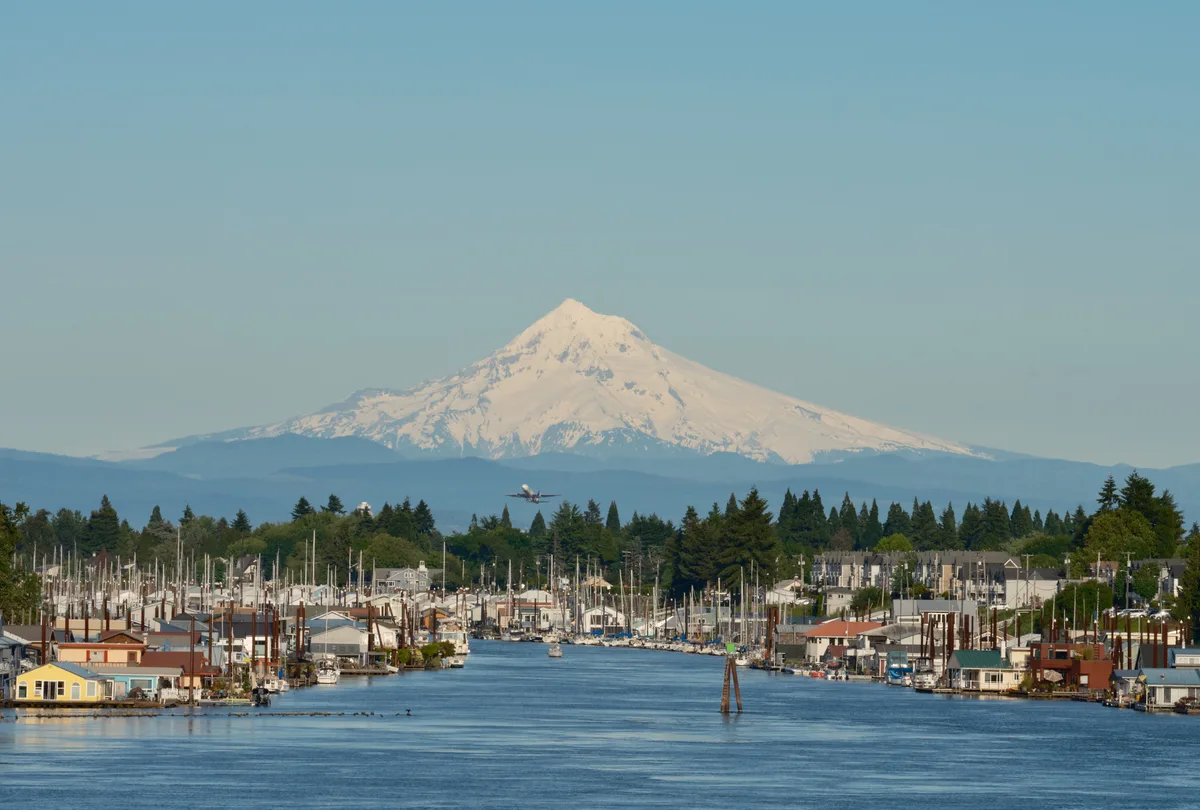 Mount Hood's snow-capped peak dominates the distant horizon beyond Portland's North Harbor, where floating homes and marinas line the Columbia River's protected channels. A commercial aircraft climbs into the pale blue sky from nearby Portland International Airport, while the foreground reveals an intimate waterfront community of colorful houseboats and recreational vessels. The serene waterway reflects the afternoon light, creating a harmonious blend of urban aviation, maritime living, and Oregon's iconic volcanic sentinel.