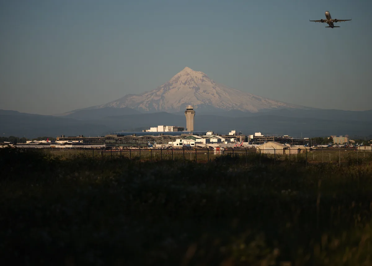 Golden hour light bathes Mount Hood's snow-capped peak as it towers majestically behind Portland International Airport's sprawling terminals and iconic control tower. A commercial airliner cuts across the hazy sky in the upper right frame, while the airport's geometric infrastructure creates strong horizontal lines against the mountain's dramatic vertical presence. The foreground vegetation frames the scene in deep shadows, emphasizing the luminous quality of the distant volcanic summit.