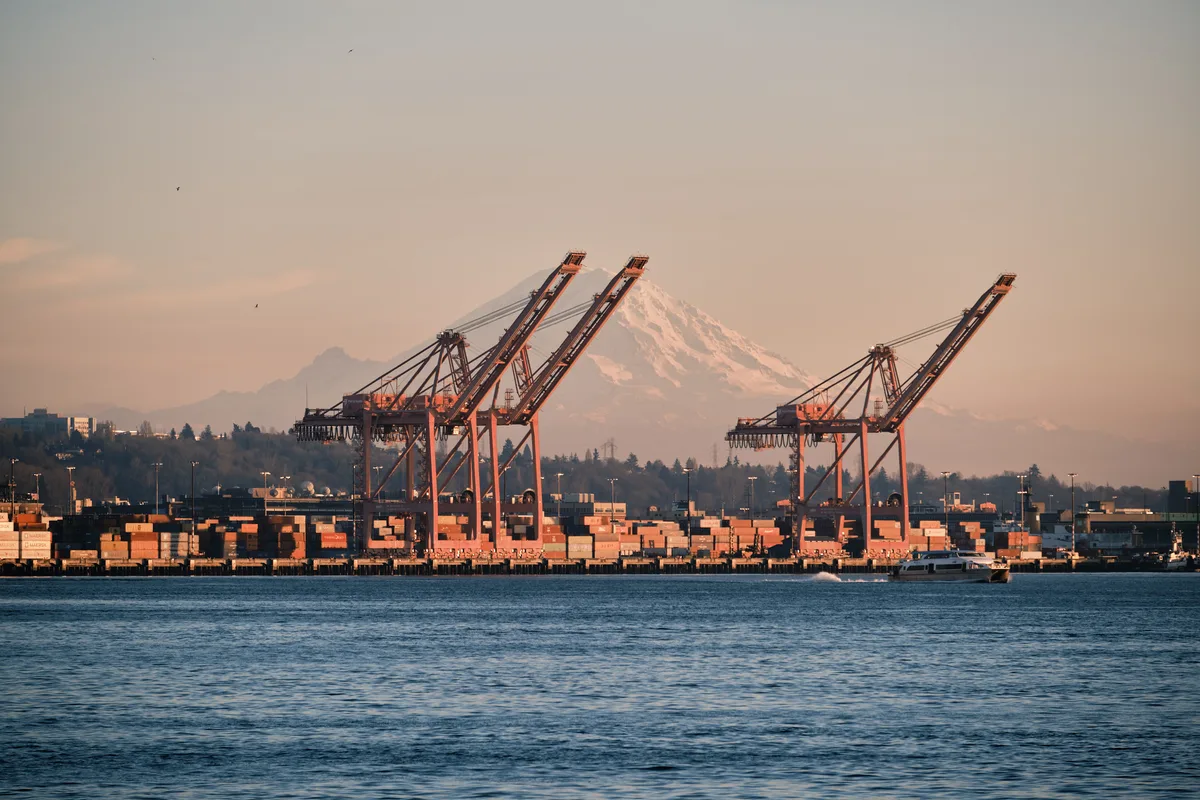 The majestic snow-capped peak of Mount Rainier dominates the horizon behind Seattle's bustling container port, where towering orange gantry cranes stand sentinel over geometric stacks of cargo containers. Golden hour light bathes the industrial waterfront in warm amber tones, while gentle ripples disturb Elliott Bay's surface as a small vessel navigates the foreground. The composition creates a striking juxtaposition between Washington's natural grandeur and the relentless pulse of maritime commerce.