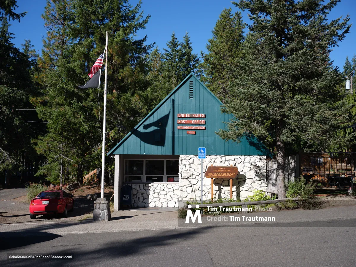 A distinctive turquoise and stone post office building nestled among towering evergreens in Government Camp, Oregon, serves as a charming community anchor beneath brilliant azure skies. The A-frame structure combines rustic stone masonry with weathered blue siding, while the American flag flies proudly against the dense Pacific Northwest forest backdrop. A red convertible idles in the foreground, adding a splash of color to this quintessential small mountain town scene bathed in golden afternoon light.