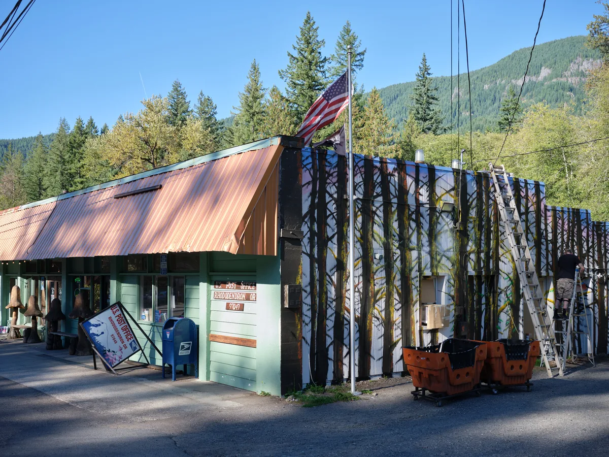 In the shadow of Oregon's Cascade Range, the weathered Rhododendron post office stands as a testament to rural American resilience. The building's patinated corrugated metal roof catches afternoon light while an American flag flies proudly above the modest mint-green structure. Towering Douglas firs frame the scene against azure skies, their ancient presence dwarfing the humble outpost that serves this mountain community along the Mount Hood Highway.