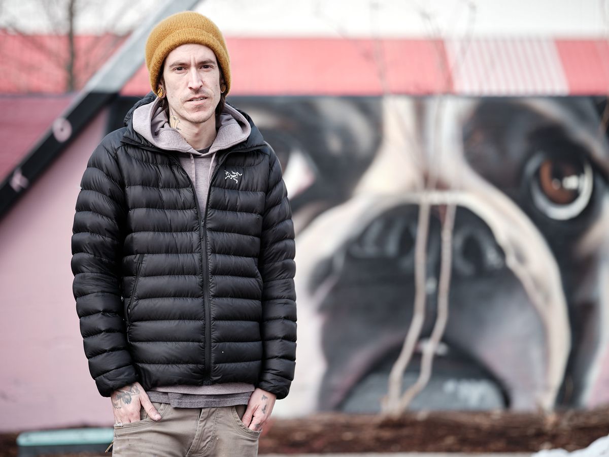 A portrait of muralist Kyle Danley, also known as Wetiko, standing in front of one of his large-scale murals depicting a close-up of a French Bulldog's face on a pink building. He wears a black Arc'teryx puffer jacket, a grey hoodie, khaki jeans, and a mustard yellow beanie. Neck and hand tattoos are visible. He looks directly into the camera with a calm, serious expression, hands in his pockets.