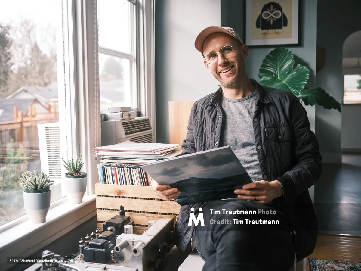 A smiling man wearing glasses and a brown cap sits by a window holding a vinyl record, surrounded by his collection of albums stored in wooden crates and vintage audio equipment. Natural light streams through the window illuminating the cozy indoor space with plants and artwork.
