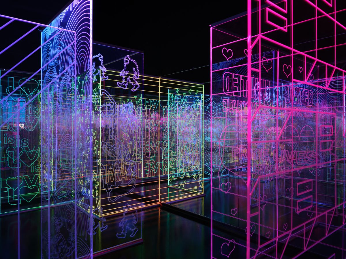 A vibrant digital art installation featuring glowing neon wireframe structures in pink, blue, and green colors against a dark background. The geometric forms include cubes, architectural elements, and text overlays creating an immersive interactive environment.