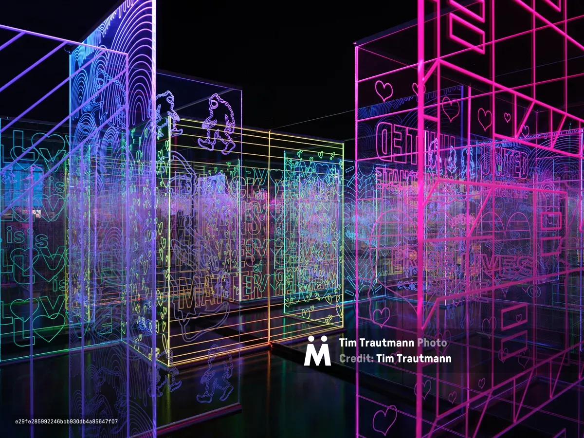 A vibrant digital art installation featuring glowing neon wireframe structures in pink, blue, and green colors against a dark background. The geometric forms include cubes, architectural elements, and text overlays creating an immersive interactive environment.