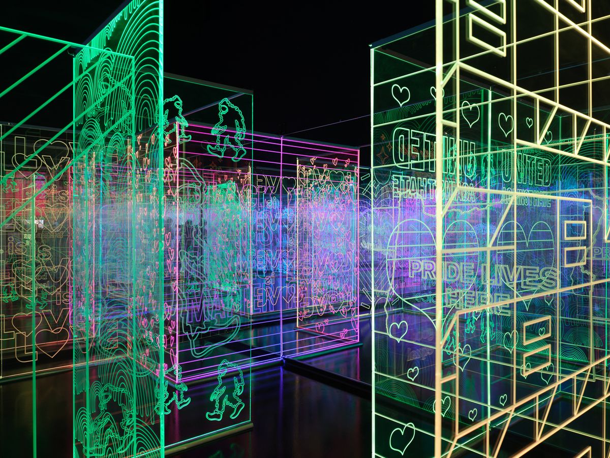 A vibrant interactive art installation featuring glowing neon outlines in green, purple, and blue colors creating geometric patterns, text, and heart shapes against a dark background. The illuminated display creates an immersive digital art experience with layered transparent panels and wireframe designs.