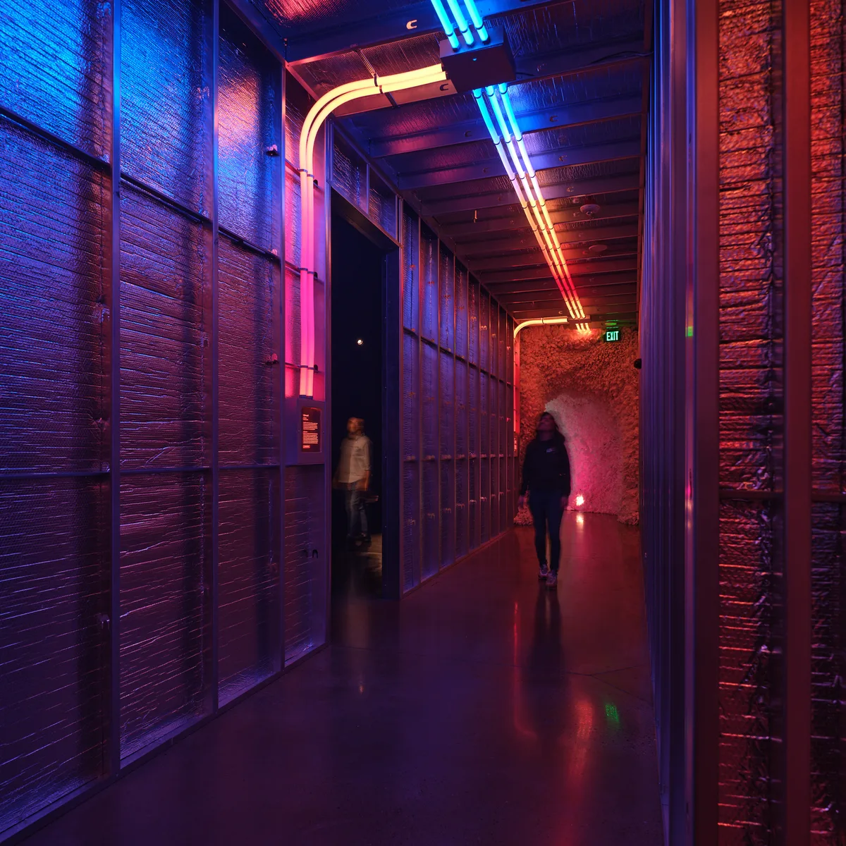 A dramatically lit hallway at Hopscotch entertainment venue in Portland, Oregon, featuring exposed brick walls illuminated by vibrant blue and red neon lighting. Two silhouetted figures walk through the atmospheric corridor with its industrial ceiling and polished concrete floors.