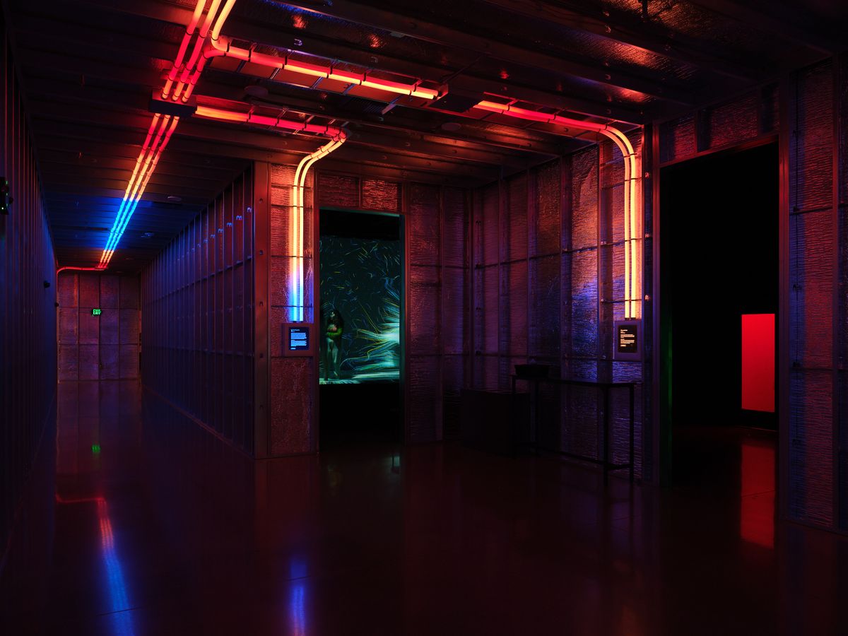 A dramatic interior hallway at Hopscotch venue in Portland featuring exposed brick walls, industrial ceiling fixtures, and vibrant neon lighting in red, blue, and green creating atmospheric reflections on the polished floor. The space showcases modern industrial design with color-changing LED tubes and digital displays along the corridor.