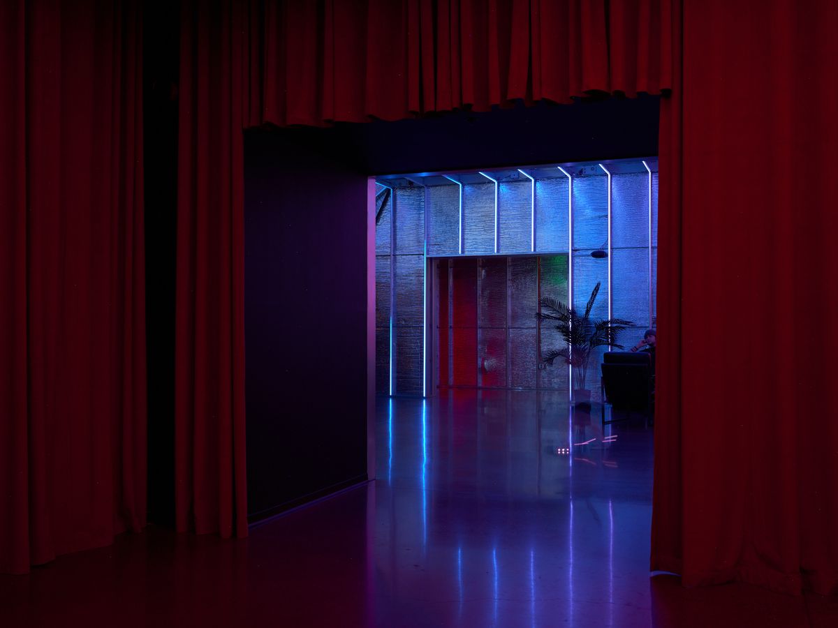 A moody interior space featuring dramatic red curtains on the left contrasting with a brightly lit modern area bathed in blue and purple neon lighting. The scene shows reflective flooring and contemporary architectural elements creating a sophisticated nightclub or entertainment venue atmosphere.