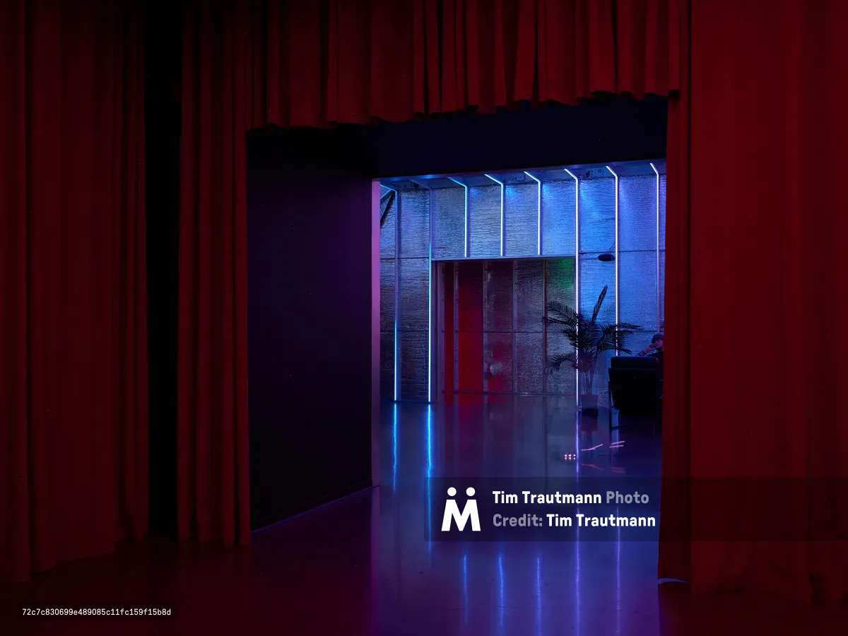 A moody interior space featuring dramatic red curtains on the left contrasting with a brightly lit modern area bathed in blue and purple neon lighting. The scene shows reflective flooring and contemporary architectural elements creating a sophisticated nightclub or entertainment venue atmosphere.