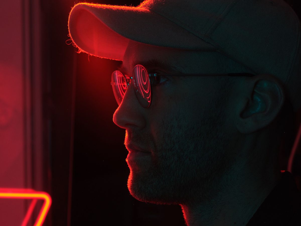 A dramatic side profile portrait of a person wearing reflective sunglasses and a hat, illuminated by vibrant red and blue neon lighting in a dark urban setting. The neon light creates striking rim lighting effects and colorful reflections in the eyewear.