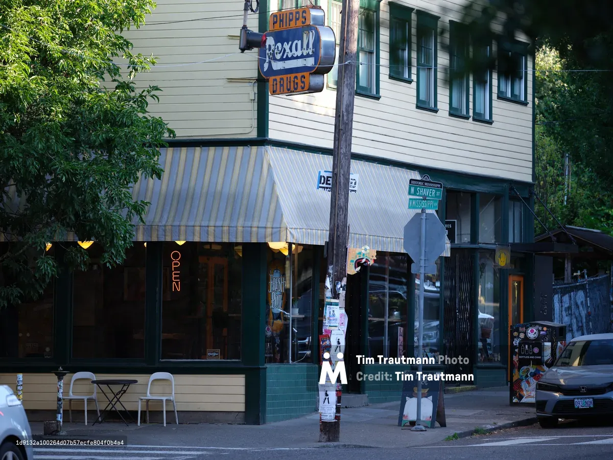 Once a Rexall Drugs pharmacy, this charming two-story building on North Mississippi Avenue now houses The Fresh Pot coffee shop. White horizontal siding, green trim, a striped awning, and an illuminated 'OPEN' sign give it a nostalgic small-town feel in the heart of historic Portland.