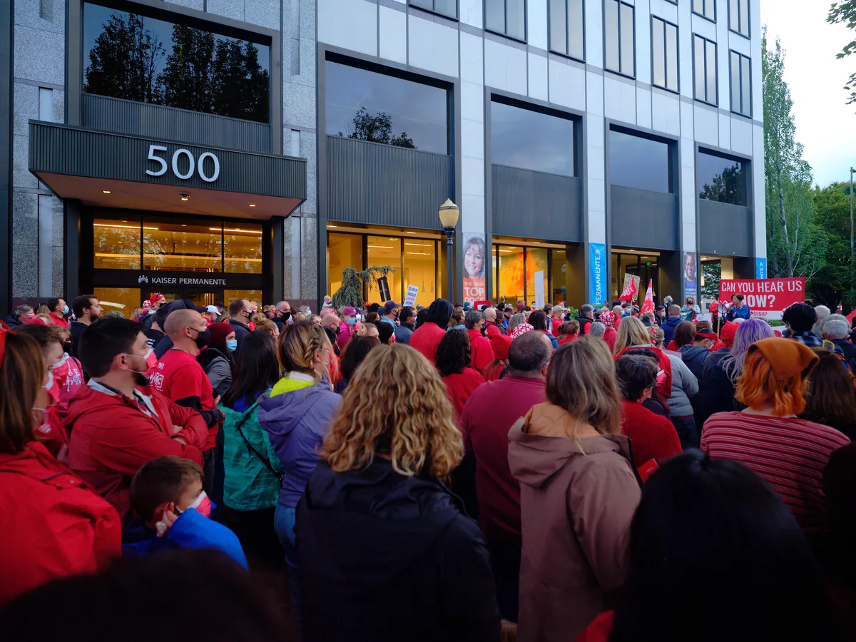 A sea of red-clad healthcare workers fills the sidewalk outside Kaiser Permanente's modernist headquarters at 500 Northeast Multnomah Street in Portland's Lloyd District. The crowd of striking nurses creates a powerful visual contrast against the building's sleek glass facade and concrete columns, their unified presence illuminated by the warm glow spilling from the medical center's ground-floor windows in the gathering dusk. Signs demanding "CAN YOU HEAR US NOW?" punctuate the peaceful but determined assembly, while the institutional architecture looms overhead, emphasizing the David-versus-Goliath nature of the labor dispute.
