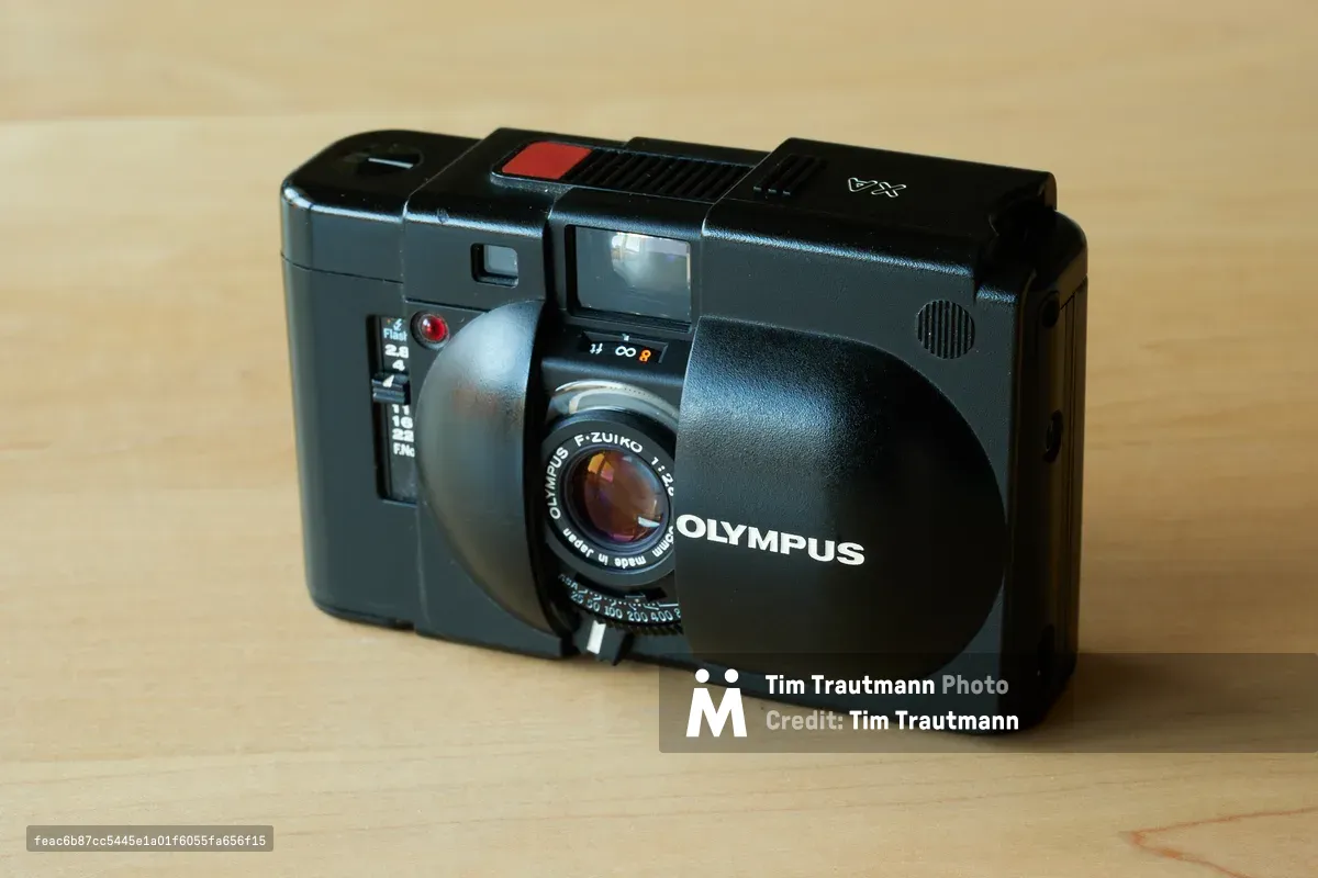 An Olympus XA compact rangefinder camera rests on warm blonde wood, its distinctive charcoal metallic body catching soft, diffused light. The legendary f/2.8 Zuiko lens peers through its protective sliding cover, while the camera's minimalist design and precision engineering speak to decades of street photography heritage. The intimate lighting creates gentle shadows that accentuate the camera's compact form and tactile surfaces.