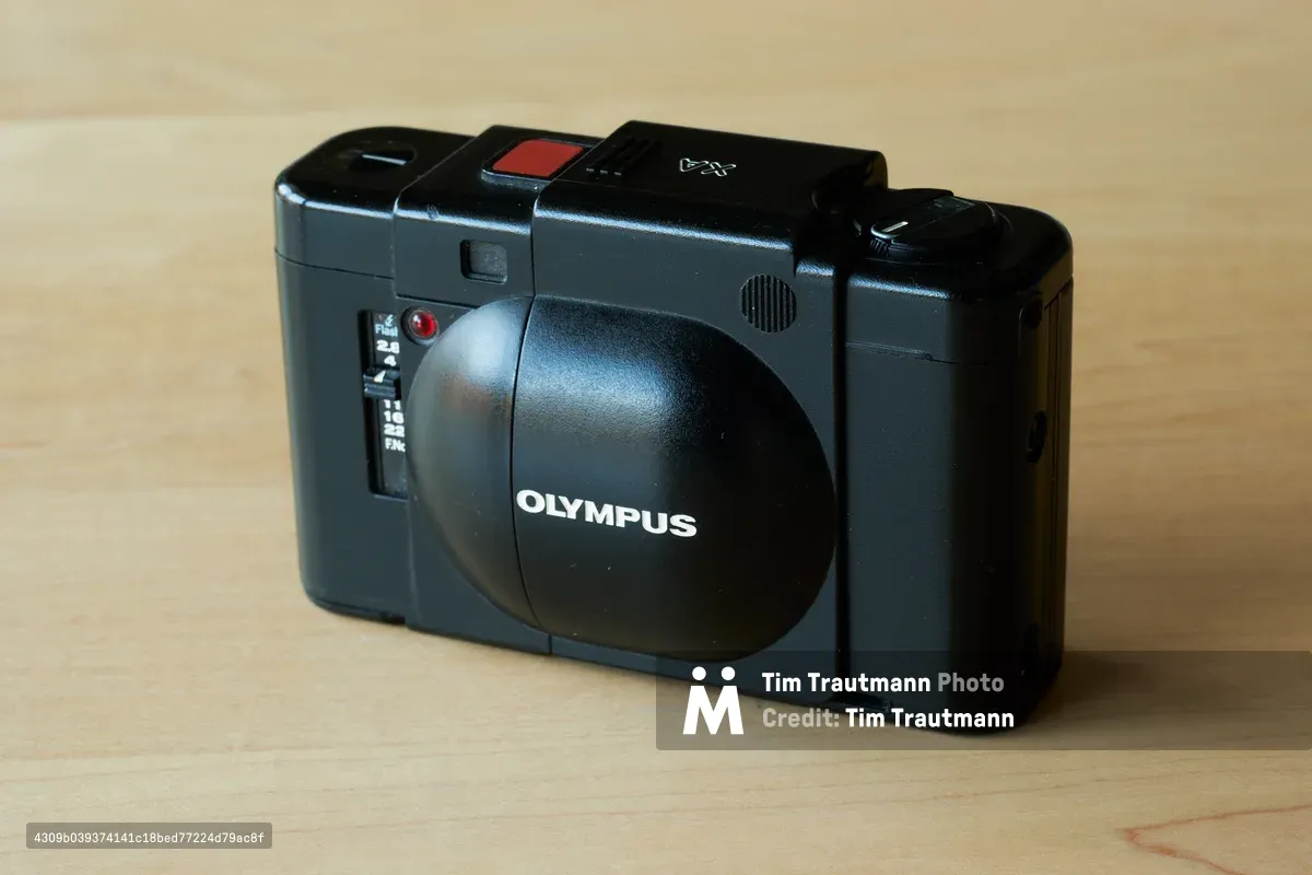 A pristine Olympus XA rangefinder camera rests on warm blonde wood, its charcoal metallic body gleaming under soft natural light. The compact 35mm camera's distinctive sliding lens cover reveals the aperture priority f/2.8 lens, while the red shutter release button provides a subtle accent against the matte black top plate. The intimate composition captures the camera at a three-quarter angle, emphasizing its legendary portable design that made it a favorite among street photographers.