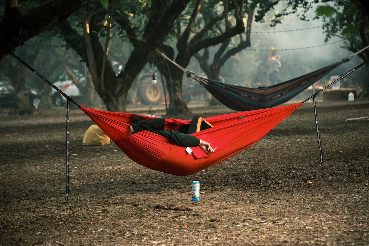 A person in black clothing reclines in a bright red hammock at the Orchard Vibration campout in Newberg, Oregon, with a canned drink resting on the ground below them. A second patterned hammock in blue and orange is visible in the background, suspended between large orchard trees. Hazy smoke drifts through the moody, dimly lit grove, with festival-goers, a tractor, string lights, and camping gear faintly visible among the trees.