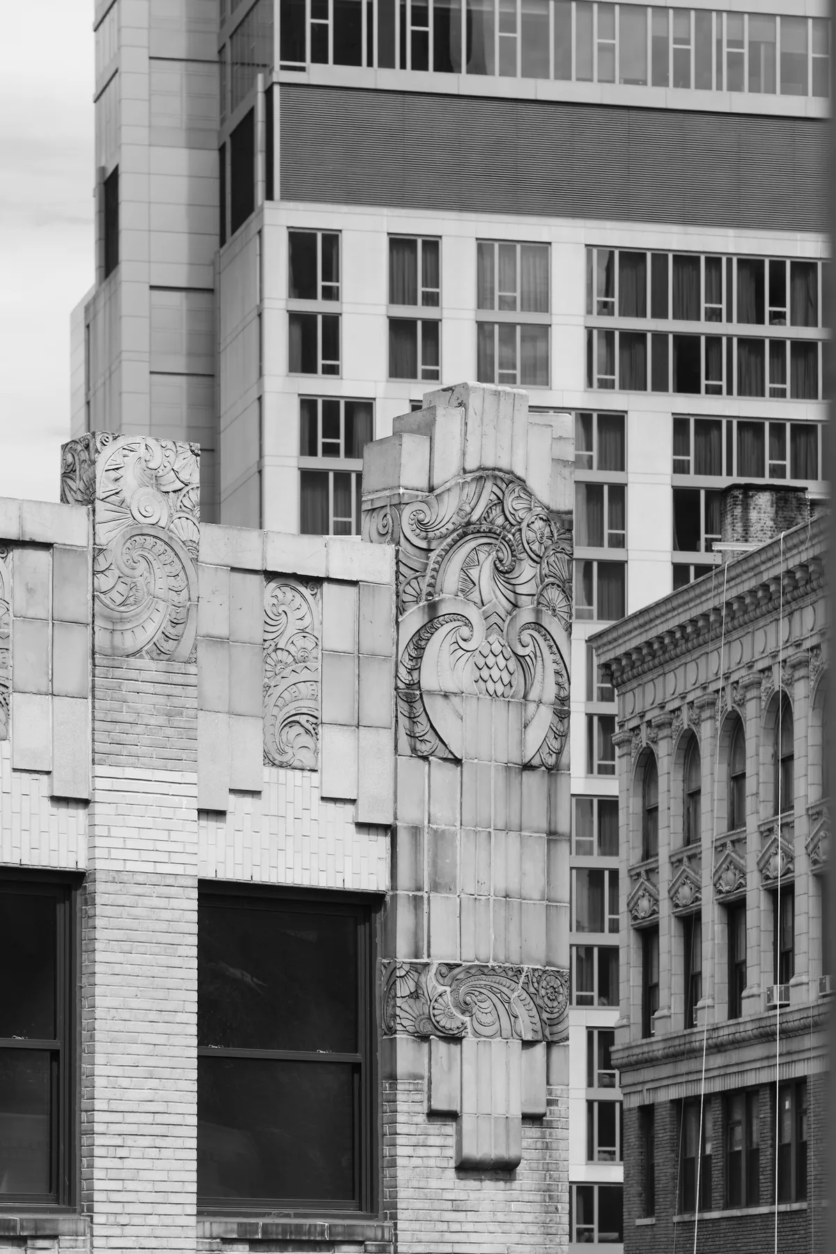 Elaborate carved stone ornaments crown the corner of a historic building in Manhattan's NoMad district, their intricate Art Deco flourishes creating dramatic shadows against weathered limestone. The monochromatic composition captures the interplay between ornate early 20th-century craftsmanship and the stark modernist glass tower rising behind, while classical arched windows peek from the right frame. Soft, diffused light emphasizes the sculptural depth of the decorative elements, revealing every carved detail in the building's ornamental crown.