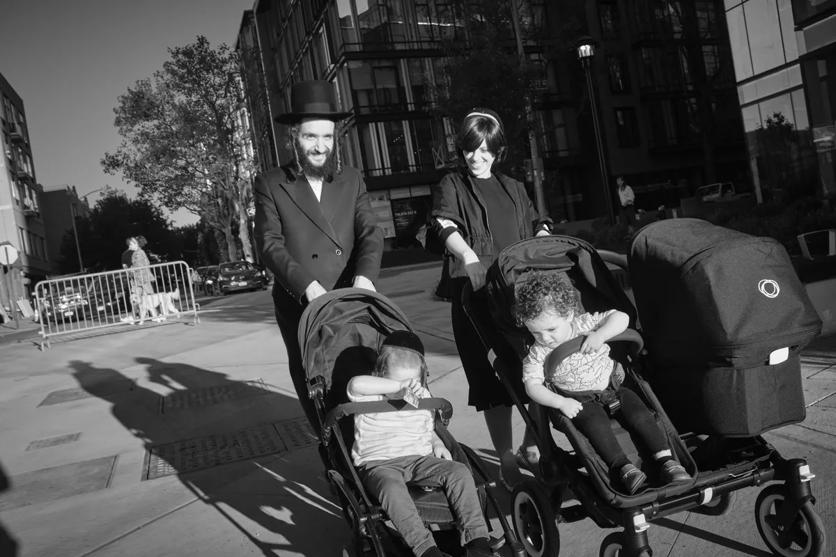 A Hasidic couple guides their double stroller through the sun-dappled sidewalks of Williamsburg, Brooklyn, their traditional black attire creating stark silhouettes against the urban landscape. The father, wearing his characteristic black hat and coat, beams with quiet joy while the mother, in modest dress and sunglasses, tends to their young children nestled in the modern stroller. Dramatic shadows stretch across the concrete as pre-war tenements with fire escapes tower overhead, capturing the intersection of timeless tradition and contemporary city life along Kent Avenue.