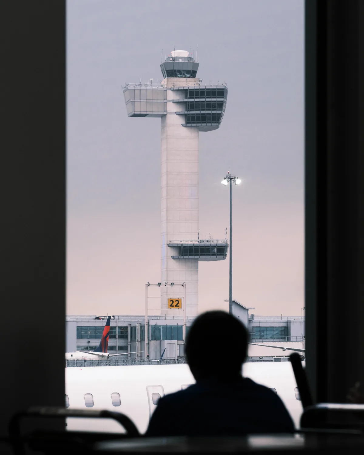 A silhouetted passenger gazes through an airport window at the iconic white control tower of John F. Kennedy International Airport, its distinctive cylindrical form crowned with a glass-enclosed observation deck. The scene captures the liminal moment of air travel, framed by the aircraft's interior in deep shadow against the tower's stark modernist architecture. Soft, diffused light creates an atmospheric haze around the control tower, while gate 22 is visible at its base, anchoring this quintessential airport tableau in Queens.